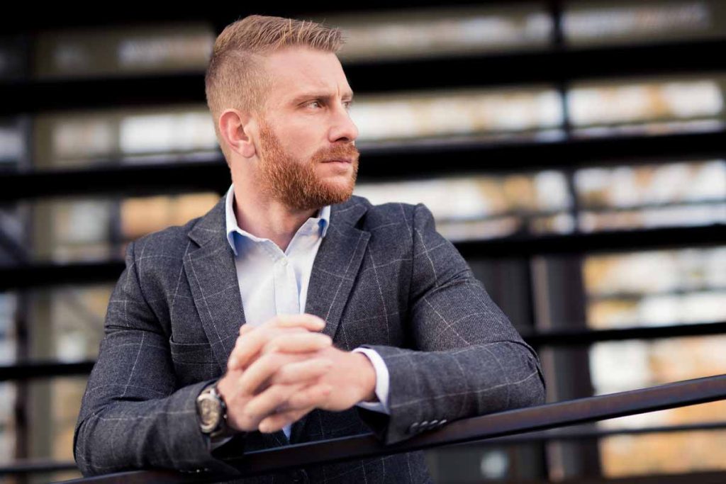 Man with a short beard wearing a suit jacket while looking off to the side outdoors.