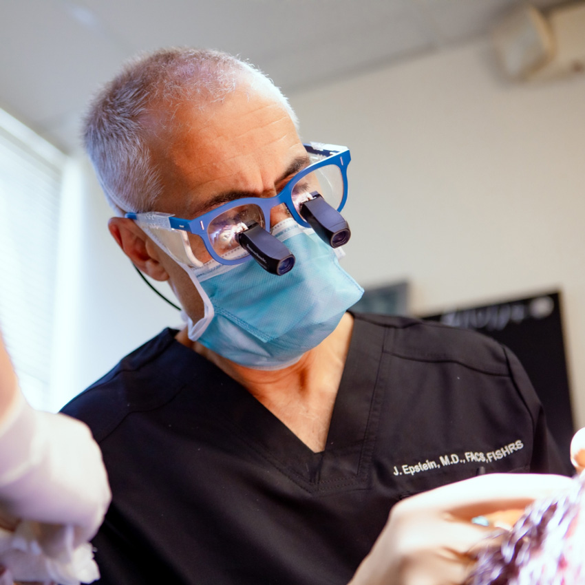 Dr. Jeffrey Epstein, Miami board-certified facial plastic surgeon, performing a surgical procedure while wearing magnifying loupes and a face mask.