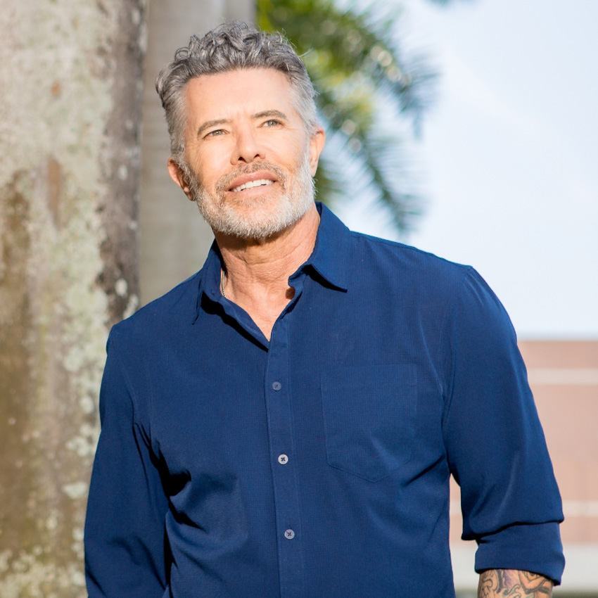 Hombre sonriente de pie al aire libre con una camisa azul abotonada.