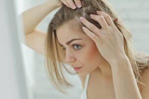 Woman examining her hairline in a mirror while parting her hair with both hands.